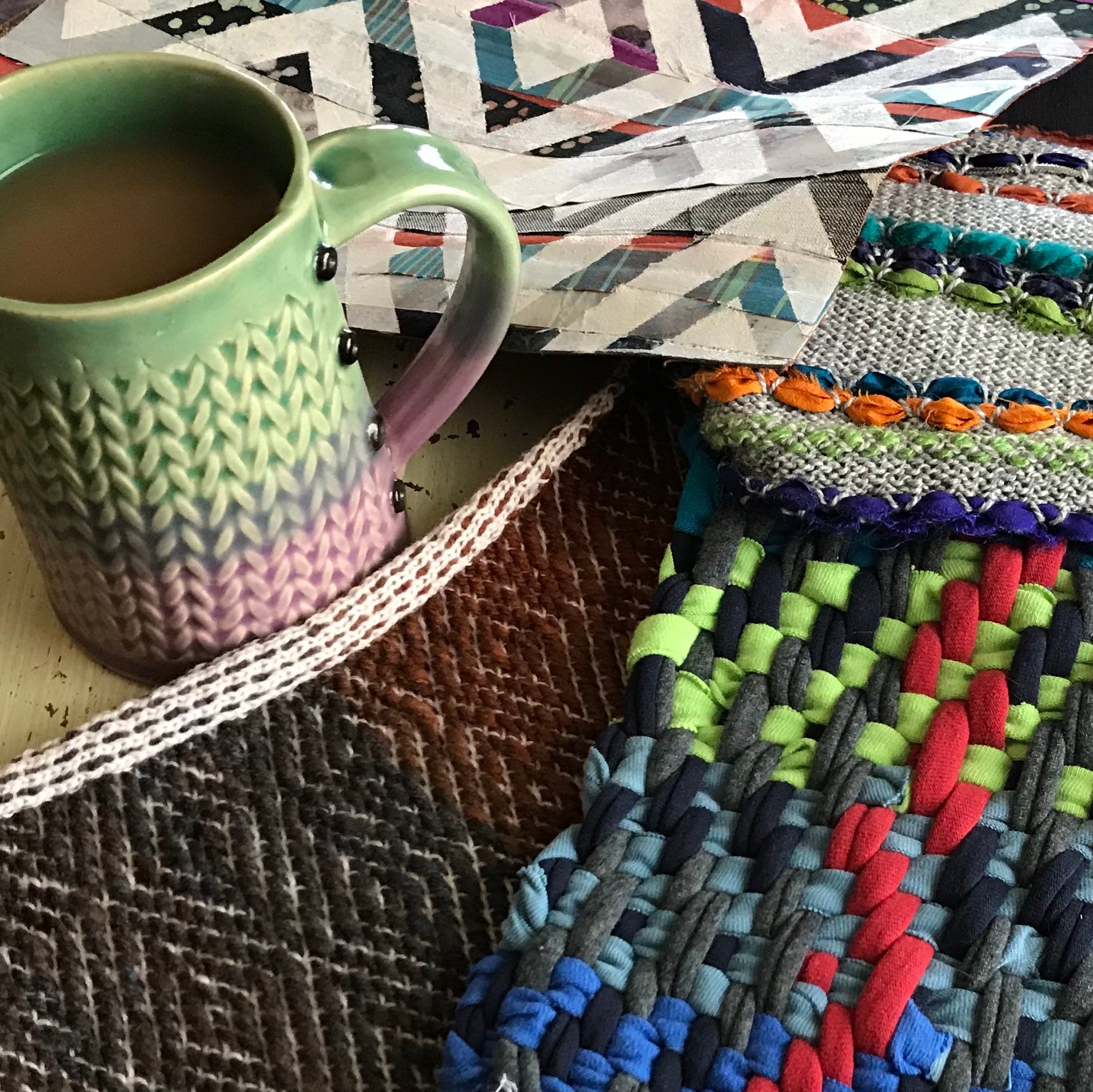 Vibrant and textured textile art samples on the desktop of designer Kathy Barlow. There is a machine knit diamonds pattern, a handwoven t-shirt yarn sample, a machine knit piece with inlaid sari silk ribbons and a layered quilted and screen-printed multimedia piece. Also on the desktop is a Charan Sachar knit-look coffee mug filled with coffee and milk.